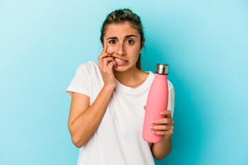 Young blonde caucasian woman holding a thermo isolated on blue background biting fingernails, nervous and very anxious.