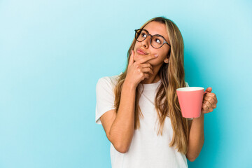 Young caucasian blonde woman holding a mug isolated on blue background looking sideways with doubtful and skeptical expression.