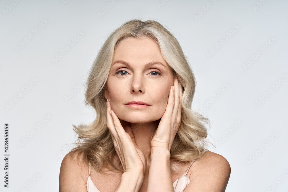 Wall mural Adult senior older woman touching her perfect skin. Beautiful portrait mid 50s aged woman advertising facial anti age lift products salon care tighten skin isolated on white looking at camera.