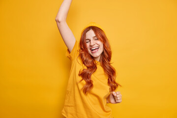 Fototapeta premium Carefree positive young ginger girl dances happily keeps arm raised closes eyes feels upbeat dressed in casual t shirt and hat poses against vivid yellow background has fun enjoys successful day