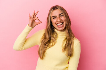 Young blonde caucasian woman isolated on pink background winks an eye and holds an okay gesture with hand.