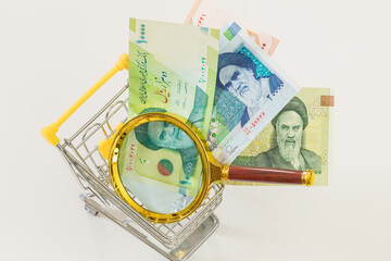 A miniature shopping cart with Iranian currency and a magnifying glass. Economic and business concept, rising inflation and the country's economic upset. White background