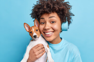 Photo of joyful beautiful Afro American girl has curly hair smiles broadly poses with pedigree dog enjoys spending free time while listening music isolated over blue background. Happy pet owner