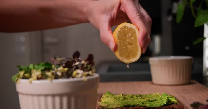 Close up panning shot of woman's hand squeezing lemon juice on avocado spread on bread, making vegan sandwich with avocado and microgreens, healthy eating concept. 