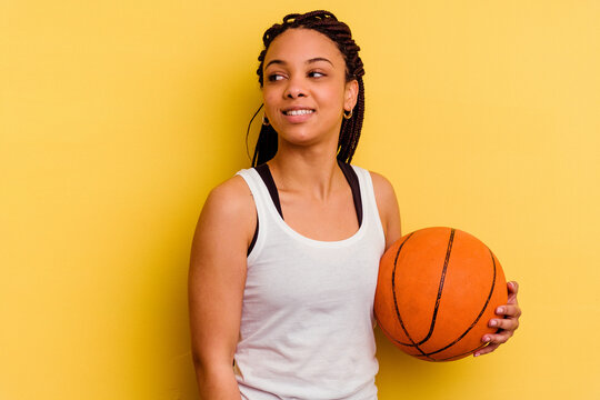 Young african american woman playing basketball isolated on yellow background looks aside smiling, cheerful and pleasant.