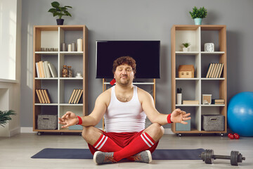 Overweight fat millennial man in sportswear with eyes closed relaxing on lotus position meditating calm down practicing yoga sitting on mat at living room. Home exercise, sports activity on quarantine - obrazy, fototapety, plakaty