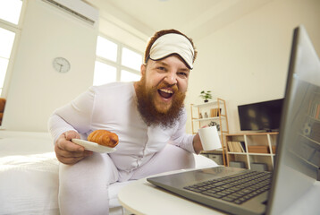 Happy fat young man indulging in food and having fun in lockdown. Portrait of plump guy in pajamas sitting on bed with laptop on Sunday morning, laughing, eating breakfast, enjoying coffee and pastry