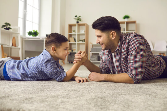 Happy boy and his dad spending time at home and having fun together. Side view of father and son lying on floor, looking at each other and arm wrestling. Family relationship, parent and child concept