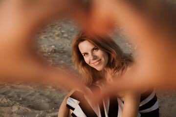 Young woman sits on the beach in the rays of sunset and smile. Child shows a smiling mother a heart by hands gesture, symbol of love. Relationship parents and children. Mother day. Family confidence