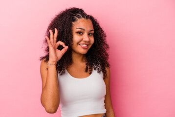 Young african american woman isolated on pink background winks an eye and holds an okay gesture with hand.