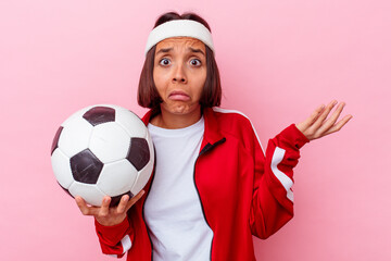 Young mixed race woman playing soccer isolated on pink background shrugs shoulders and open eyes...