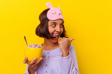 Young mixed race woman eating cereals wearing a pijama isolated on yellow background points with...