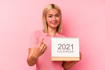 Young venezuelan woman holding a calendar isolated on pink background pointing with finger at you as if inviting come closer.