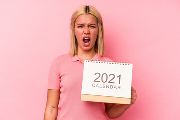 Young venezuelan woman holding a calendar isolated on pink background screaming very angry and aggressive.