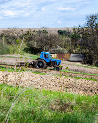 old blue tractor with plow on field and cultivates soil on background of hilly landscape in Ukrainian village. Preparing the soil for planting vegetables in spring. Agricultural machinery, field work.