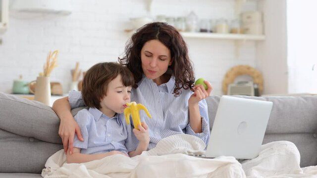 Smiling mother with little son watch funny video, cartoon on laptop computer sit together on sofa in living room. Mom and cute boy with apple and banana fruit under blanket spend time at funny movie