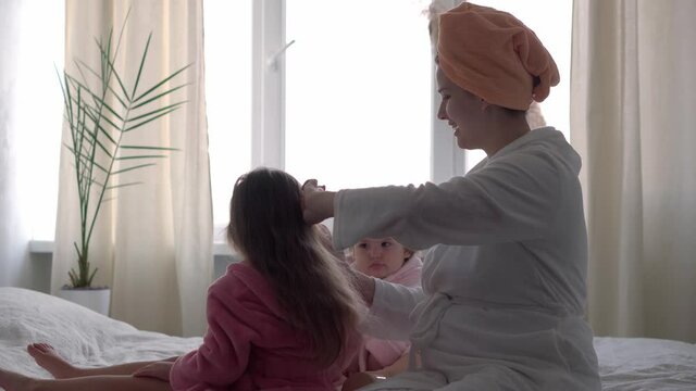 Authentic cute young woman combing long hair of baby girl. mom with 2 children daughters different ages in bathrobes after bath on white bed oposite window. Spa, motherhood, childhood, family concept.