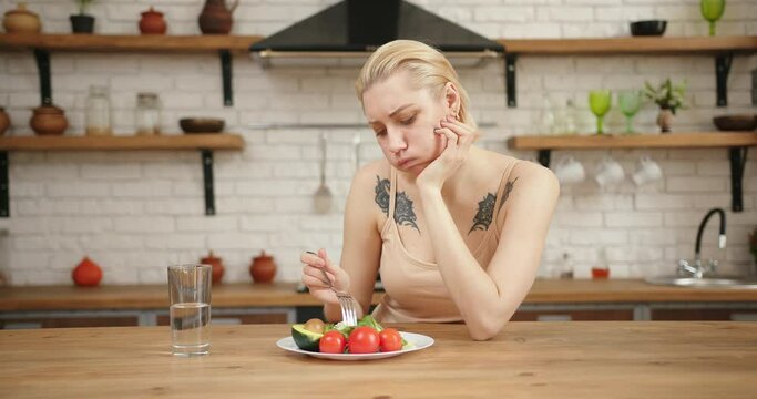 Caucasian blond woman with acne sitting at table, looking sad and bored with diet not wanting to eat salad. Modern kitchen interior