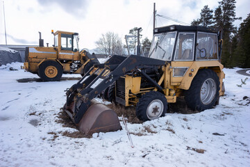 Two old rusty loader tractors stand on a snow ground - small and big, winter time.