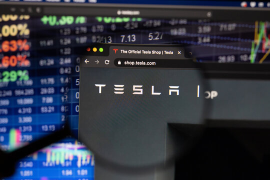 Tesla company logo on a website with blurry stock market developments in the background, seen on a computer screen through a magnifying glass