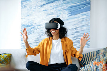 Excited african american carefree girl, sitting on a sofa at home and gesturing hands while testing new app via VR device. Cheerful curly modern girl using VR glasses, virtual reality concept