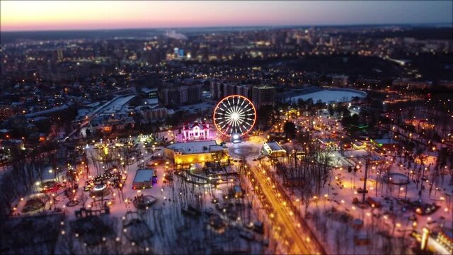  Aerial view on winter Kharkiv city center in sunset, amusement Central Park with Ferris wheel drone left to right. City lights, streets with blurred background effect