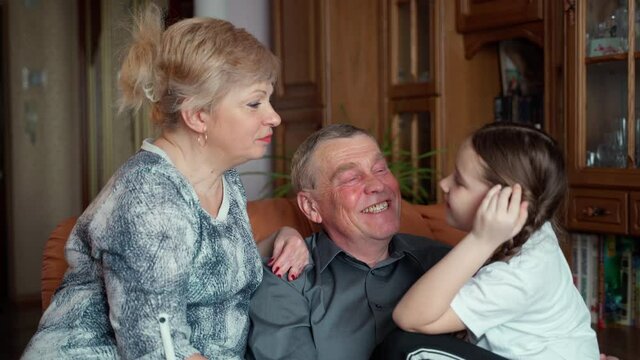 Elderly 60s grandparents and a young granddaughter, a girl, are sitting on the couch at home. They smile and laugh kissing grandfather on the cheeks. Concept of two 2 generations and heritage