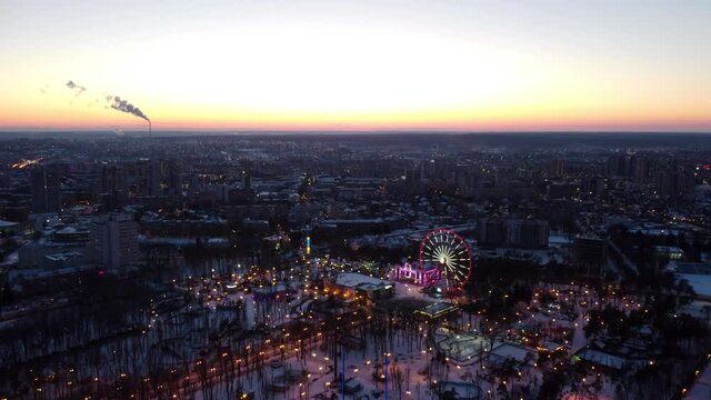 Kharkiv city center in sunset colors, amusement Gorky Central Park drone 4k panoramic left to right footage. Aerial view on winter city lights, streets and Ferris wheel entertainment