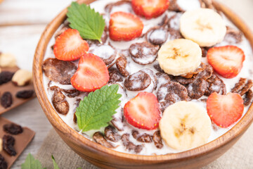 Chocolate cornflakes with milk and strawberry in wooden bowl on white wooden background. Side view, selective focus.
