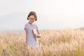 Fototapeta na wymiar Cute happy asian little girl hold book playing on meadow with sunset