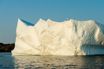 A large white iceberg formation floating in the cold ocean with layers of textured ice and snow. The ice is in transition melting from the warm rays of the sun at sunset. The berg is a blocky shape.