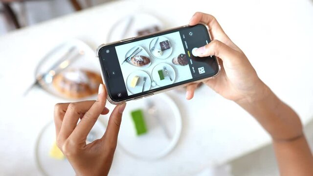 Asian woman hand using smartphone taking a photo of tasty cake dessert on white plate on the table. Female blogger influencer photographing sweet bakery and beverage for review in food restaurant blog