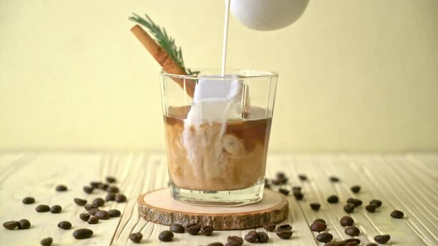 pouring milk in black coffee glass with ice cube, cinnamon and rosemary on wood background