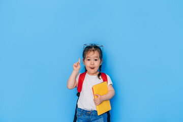 Surprised schoolgirl holding yellow book in front of blue background. Back to school in 2021....