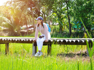 A woman carrying a blue backpack sits on a wooden bridge in the middle of a rice field sunrise effect