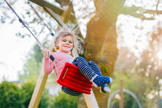 Happy beautiful little toddler girl having fun on swing in domestic garden. Cute healthy child swinging under blooming trees on sunny spring day. Baby laughing and crying