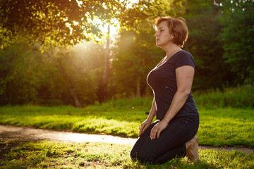 Adult woman with short hair sitting in a dark dress in the park on the grass in the rays of the setting sun summer day