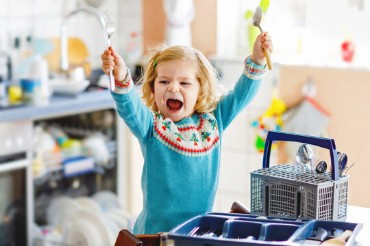 Cute little toddler girl helping in the kitchen with dish washing machine. Happy healthy blonde child sorting knives, forks, spoons, cutlery. Baby having fun with helping housework mother and father.
