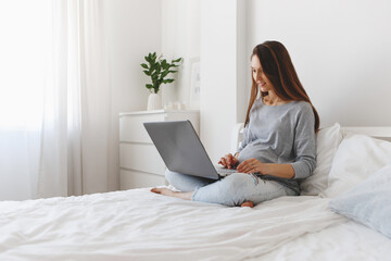 Young beautiful pregnant woman sitting on bed with laptop.
