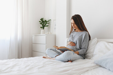 Young beautiful pregnant woman sitting on the bed and reading a book.