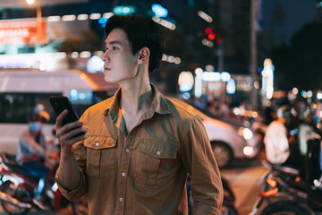 Young Asian man is using his phone while walking in the street at night