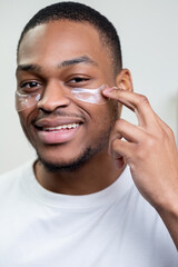 Man facial care. Skin rejuvenation. Cosmetic product advertising. Satisfied African guy in white t-shirt applying collagen gel face eye patches smiling on light background.