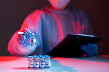Vaccine testing. Covid-19 inoculation. Pharmaceutical research. Female doctor in ppe with clipboard checking vial dose in red blue neon light on dark blur background.