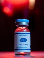 Coronavirus vaccine. Pandemic immunization. Flu inoculation. Covid-19 prevention. Dose vial glass bottle with blue label on black background with red pink neon bokeh light.