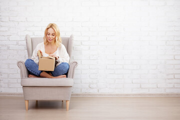 portrait of young beautiful woman sitting in armchair and opening parcel box - copy space over...
