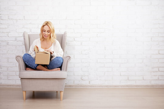 portrait of young beautiful woman sitting in armchair and opening parcel box - copy space over white brick wall