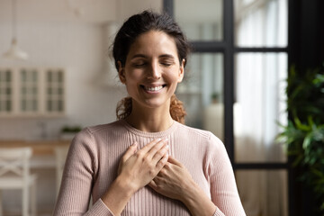 Thank you my God. Happy peaceful millennial hispanic lady stand alone with closed eyes keep hands...