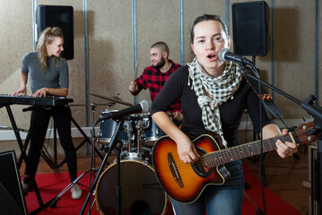 Fototapeta na wymiar Portrait of happy excited brunette girl rock singer with guitar during rehearsal with male drummer and female keyboardist in studio