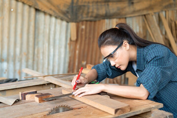 Young beautiful Asian female carpenter in blue shirt wearing safety goggles for woodworking using a...