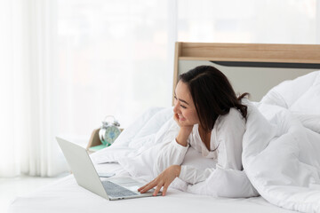 Obraz premium Young Asian female laying on bed using notebook computer. Happy female using laptop at home in bedroom. Working from home in quarantine lockdown. Women watching movie on laptop lying in bed.